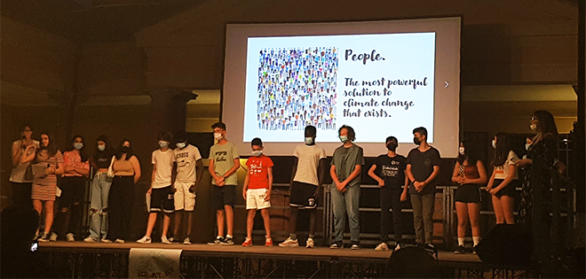 Student in a row on a stage, with face masks. A screen behind them shows a presentation with the heading "People"